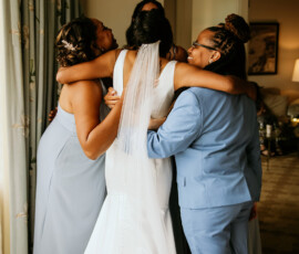 Bride in white with three women in blue dresses or suit share a warm, supportive group hug at a Gulf South wedding indoors.