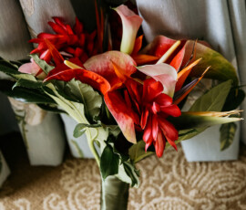 Tropical bouquet with red and pink blooms, green leaves, and ribbon on beige carpet by blue curtains—ideal for a New Orleans wedding.