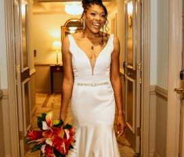 Smiling bride in a white wedding dress holds red and white bouquet in softly lit hallway before her Louisiana ceremony.
