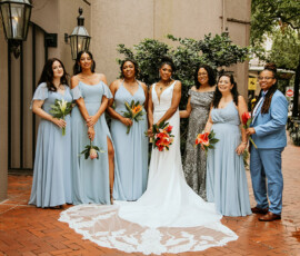 Bride in white with six attendants in blue and gray, holding tropical flowers on a brick walkway, New Orleans wedding style.