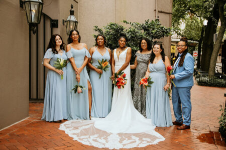 Bride in a sleek white wedding gown with a long lace train stands with her bridesmaids and wedding party in light blue dresses and a blue suit, each holding tropical bouquets, posing together on a brick walkway in New Orleans.