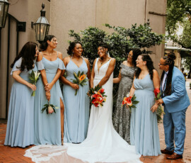 Bride in white with bridesmaids in blue and one in a blue suit, all holding tropical bouquets on a brick path by lush greenery.