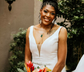 Smiling bride in a white dress with tropical bouquet, elegant updo, and necklace, greenery and lantern behind—New Orleans wedding.