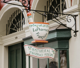 A mortar-and-pestle sign for La Pharmacie Française and Historical Pharmacy Museum hangs outside arched windows with green trim.
