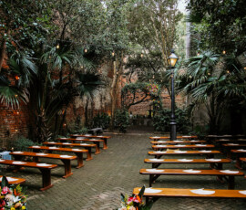 Charming Gulf South wedding courtyard with wooden benches, floral decor, brick floor, string lights, trees, and a vintage lamp post.