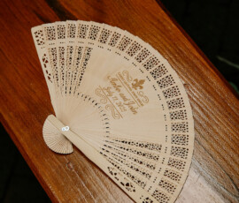 Engraved wooden hand fan with cutout designs and cursive writing on a polished bench at a Louisiana wedding reception.