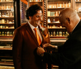 A smiling man in a rust suit gets his jacket adjusted in an old apothecary before a New Orleans wedding.