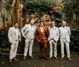Five men in suits, one in rust and four in beige, pose by florals at a Louisiana wedding, greenery and brick walls behind them.