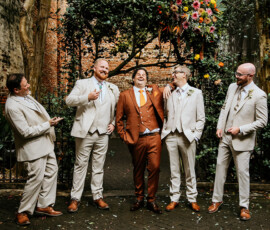 Five men, four in beige suits and one in rust, laugh under a flower arch at a Louisiana wedding with brick and greenery.
