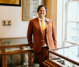 Man in rust suit with yellow tie and boutonniere stands by glass display cases in vintage museum room with wood floors.