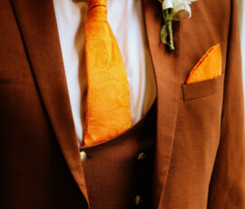 Man in brown suit with orange paisley tie and boutonnière stands by window, ready for Louisiana wedding.