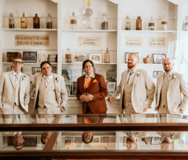Groomsmen in beige and brown suits pose by vintage pharmacy shelves in a museum, inspiring timeless Gulf South weddings.