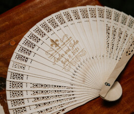 Engraved wooden hand fan with “Andrea and John July 4, 2015” in gold, open on wood surface; wedding keepsake with cutout patterns.