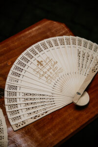 Here’s SEO-friendly alt text for this detail shot: **Alt Text:** Custom laser-cut wedding fan with Amber and John’s names and July 11, 2025 date displayed on a wooden table as a ceremony favor.