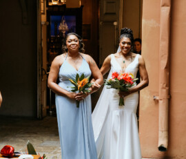 Two women in white and blue dresses smile outdoors with bouquets, surrounded by greenery and orange flowers at a rustic Louisiana wedding.