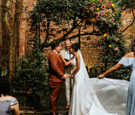 Bride and groom hold hands and smile under a floral arch at an outdoor Louisiana wedding, with veil adjustment and officiant nearby.