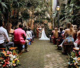 A couple exchanges vows under trees at an outdoor Louisiana wedding, with guests on benches and colorful flowers along the aisle.