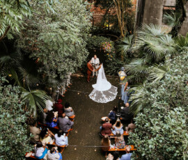Bride and groom at an outdoor wedding surrounded by greenery, with guests on benches, viewed from above.