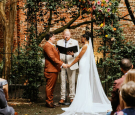 A couple holds hands under a floral arch at their Gulf South outdoor wedding as guests watch and an officiant reads.
