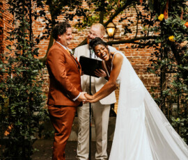 Bride and groom laugh holding hands under a floral arch at their outdoor New Orleans wedding, brick wall in background.