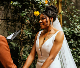 Bride in white dress holds partner’s hand at outdoor Louisiana wedding, smiling amid greenery and yellow flowers.