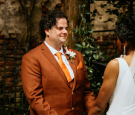 Smiling man in rust suit holds hands with woman in white dress at outdoor New Orleans wedding by rustic wall and greenery.