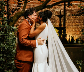 Bride in white dress and veil kisses groom in a lush garden with brick wall, timeless Louisiana wedding inspiration.