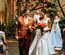 Bride and groom walk smiling down an outdoor aisle as guests watch, with petals, floral arch, and string lights in New Orleans.