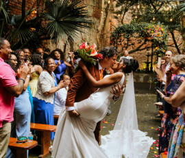 Newlyweds share a joyful kiss outdoors as guests applaud, surrounded by greenery, flowers, and hanging lights.