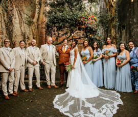 Bride, groom, and wedding party pose in a New Orleans courtyard with blue dresses, beige suits, flowers, and greenery.