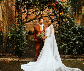 Bride in a lace gown and groom in brown suit pose under floral arch at a romantic Louisiana wedding on a cobblestone path.