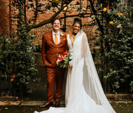 Bride in white gown and veil with groom in rust suit under floral arch, smiling outside by brick wall at Louisiana wedding.
