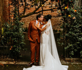 Bride and groom kiss under a floral arch in a garden wedding, with lush greenery and brick wall in the background.
