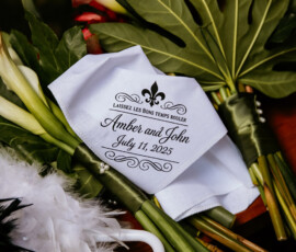 White napkin with “Amber and John, July 11, 2025,” fleur-de-lis and “Laissez les bons temps rouler” on green leaves.