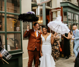 Smiling wedding couple with feathered umbrellas outside New Orleans Historic Pharmacy Museum, surrounded by guests.