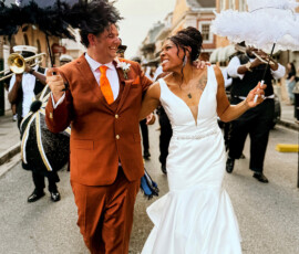 Smiling bride and groom parade with decorated umbrellas at a lively Louisiana wedding, followed by musicians and guests.