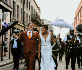 Joyful bride and groom walk a Louisiana city street with brass band and friends, celebrating their wedding with music and dance.