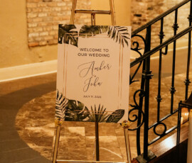Gold easel with tropical wedding welcome sign for Amber & John, July 11, 2025, indoors by brick wall and black railing.