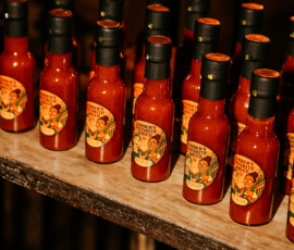 Rows of hot sauce bottles with orange labels on a wooden shelf, warm light highlighting their vibrant red color.