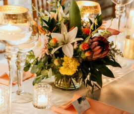 Elegant floral centerpiece with lilies on a white table, peach napkins, candles, and glass holders for Louisiana wedding inspiration.