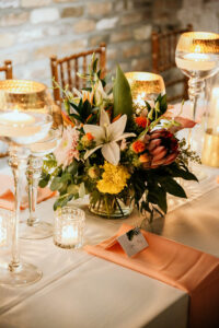 Elegant tropical wedding centerpiece with lilies, protea, marigolds, and greenery surrounded by glowing candles on a reception table with peach napkins at a New Orleans wedding celebration.
