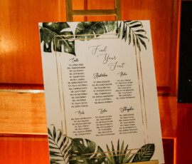 Tropical leaf seating chart on an easel with a rustic background, ideal for Gulf South wedding inspiration.