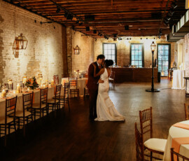 Bride and groom share a romantic kiss dancing in a warmly lit, rustic New Orleans wedding hall with wooden beams and brick walls.