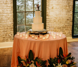Elegant white tiered wedding cake with topper on a pink table, surrounded by candles and florals at a Gulf South reception.
