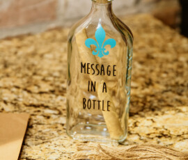 Glass bottle with blue fleur-de-lis, “Message in a Bottle,” rolled paper inside, on granite next to twine, marker, and scroll.