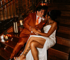 Bride in white and groom in orange suit sit on wooden stairs, holding hands amid candlelight and sharing a joyful smile.