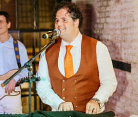 Man in brown suit and orange tie plays keyboard and sings; guitarist stands behind. Indoor brick wall and large window setting.