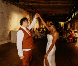 Couple dances at Gulf South wedding; man in rust suit, woman in white dress. Guests watch from tables in warmly lit venue.