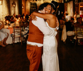 A joyful couple slow dances in formal wear at a warmly lit New Orleans wedding, surrounded by seated guests.
