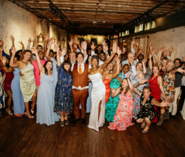 Wedding guests celebrate indoors with the bride in white and groom in brown, in a rustic hall with brick and wood floors.